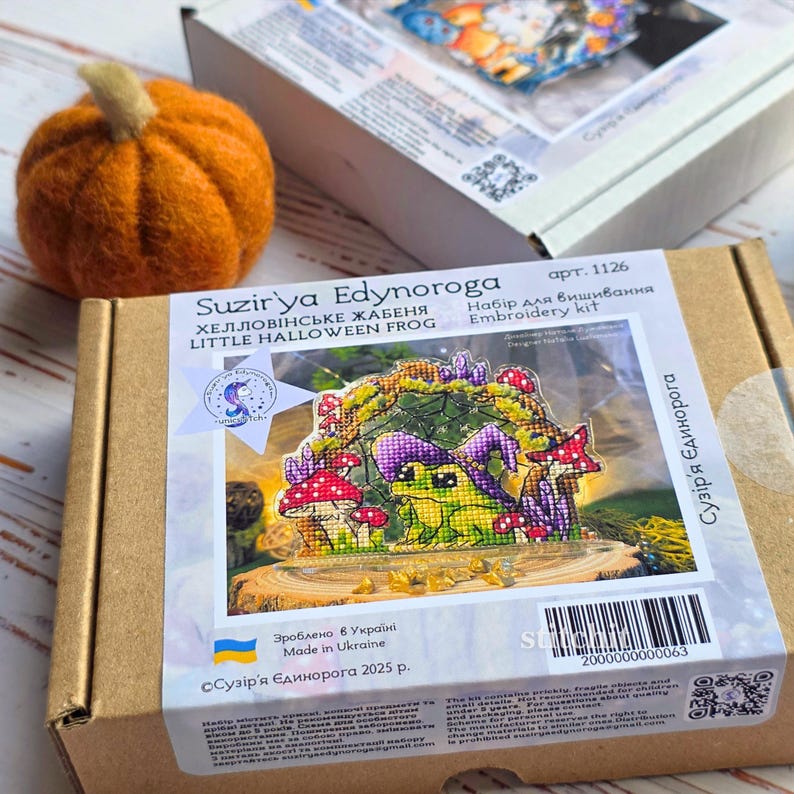 Halloween Frog Cross Stitch Kit — box front showing finished frog on mushroom design, styled with pumpkin on wooden surface