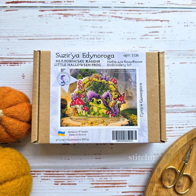 Halloween Frog Cross Stitch Kit — box packaging with finished frog in witchy hat among mushrooms and autumn leaves, styled with pumpkin and scissors