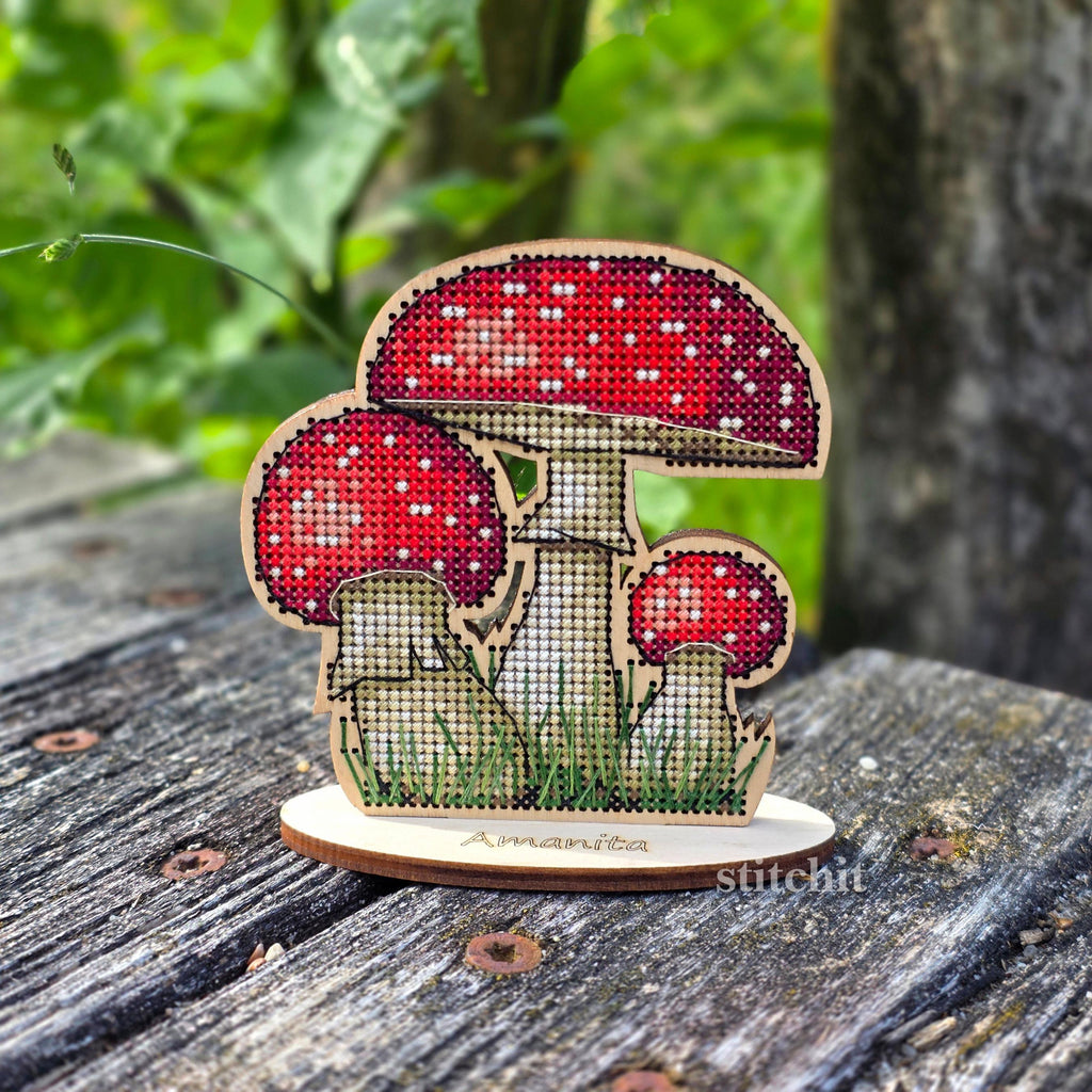 Mushroom Cross Stitch Kit on Wood — finished red fly agaric with three mushrooms and green grass on a wooden stand, displayed on a wooden surface.