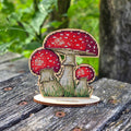 Mushroom Cross Stitch Kit on Wood — finished red fly agaric with three mushrooms and green grass on a wooden stand, displayed on a wooden surface.