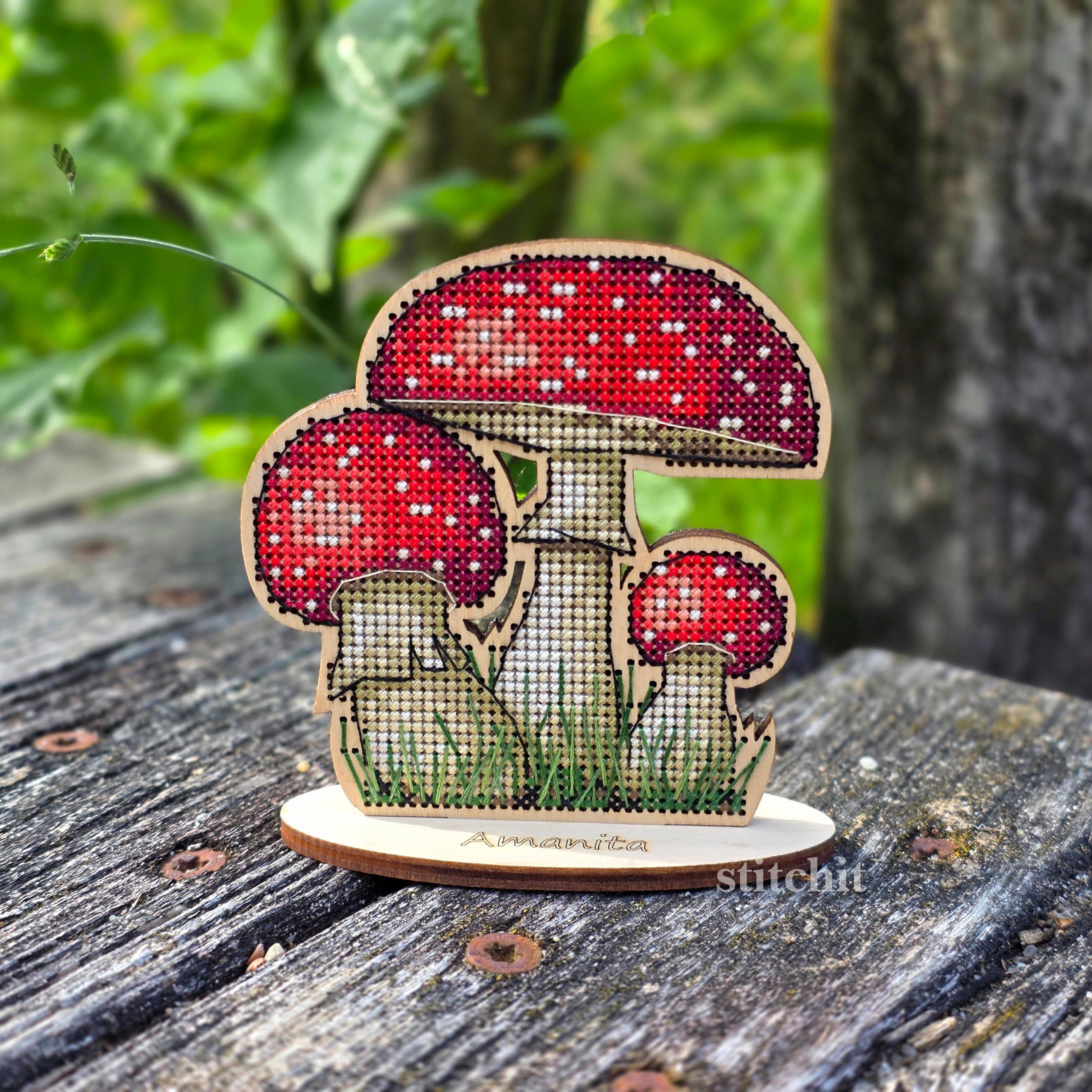 Mushroom Cross Stitch Kit on Wood — finished red fly agaric with three mushrooms and green grass on a wooden stand, displayed on a wooden surface.