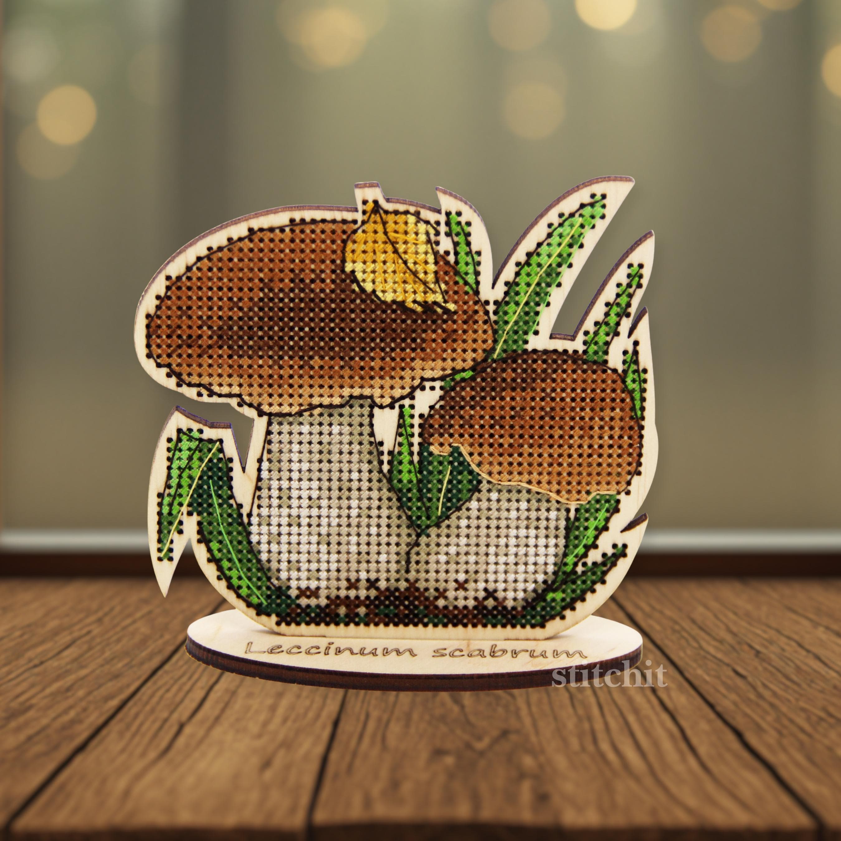 Forest Mushroom Cross Stitch Kit on Wood — finished birch bolete with green leaves and a yellow leaf on a wooden stand, standing upright on a wooden surface