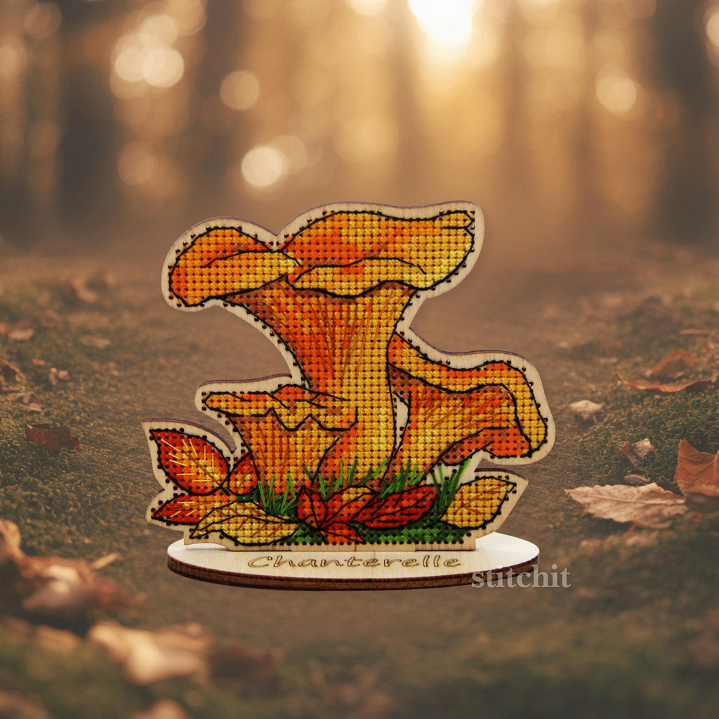 Chanterelle Cross Stitch Kit on Wood — finished golden chanterelle on a wooden stand with green grass stitching, photographed outdoors on a forest floor with fallen leaves.