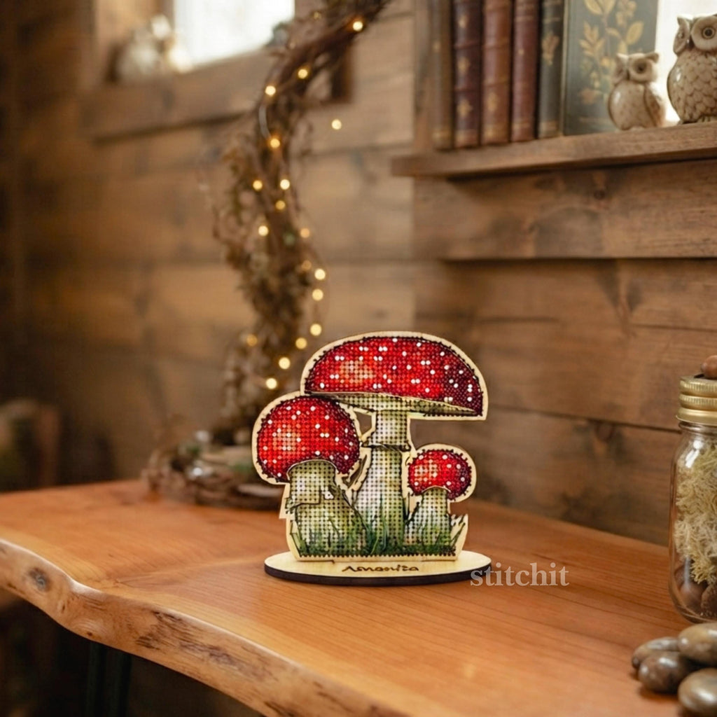 Mushroom Cross Stitch Kit on Wood — finished fly agaric on a wooden stand displayed indoors on a shelf with books, and string lights.