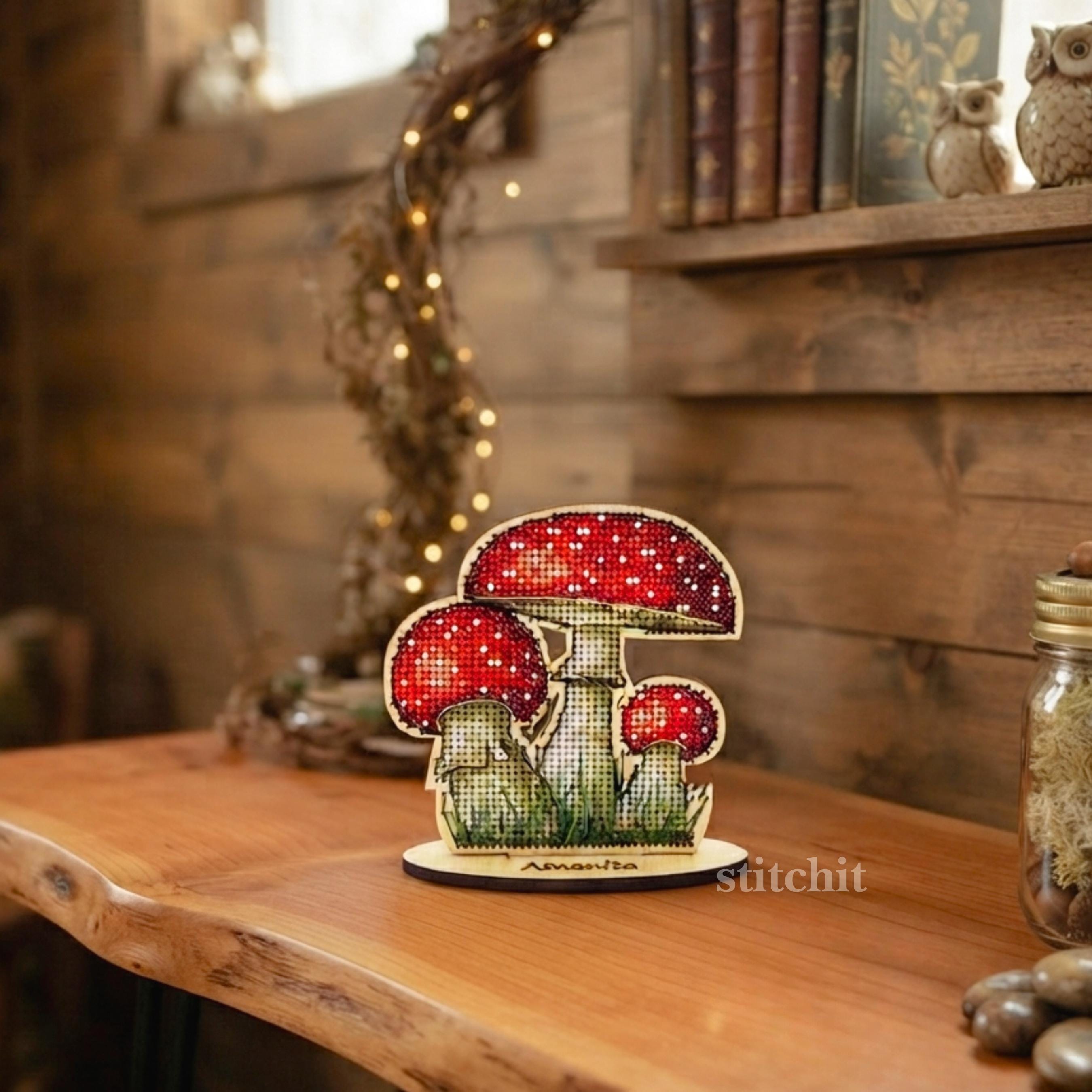 Mushroom Cross Stitch Kit on Wood — finished fly agaric on a wooden stand displayed indoors on a shelf with books, and string lights.