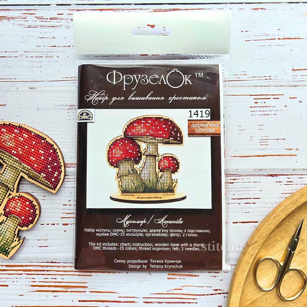 Mushroom Cross Stitch Kit on Wood — Amanita kit packaging with chart, DMC threads, and two finished fly agaric mushrooms with scissors on a white surface.