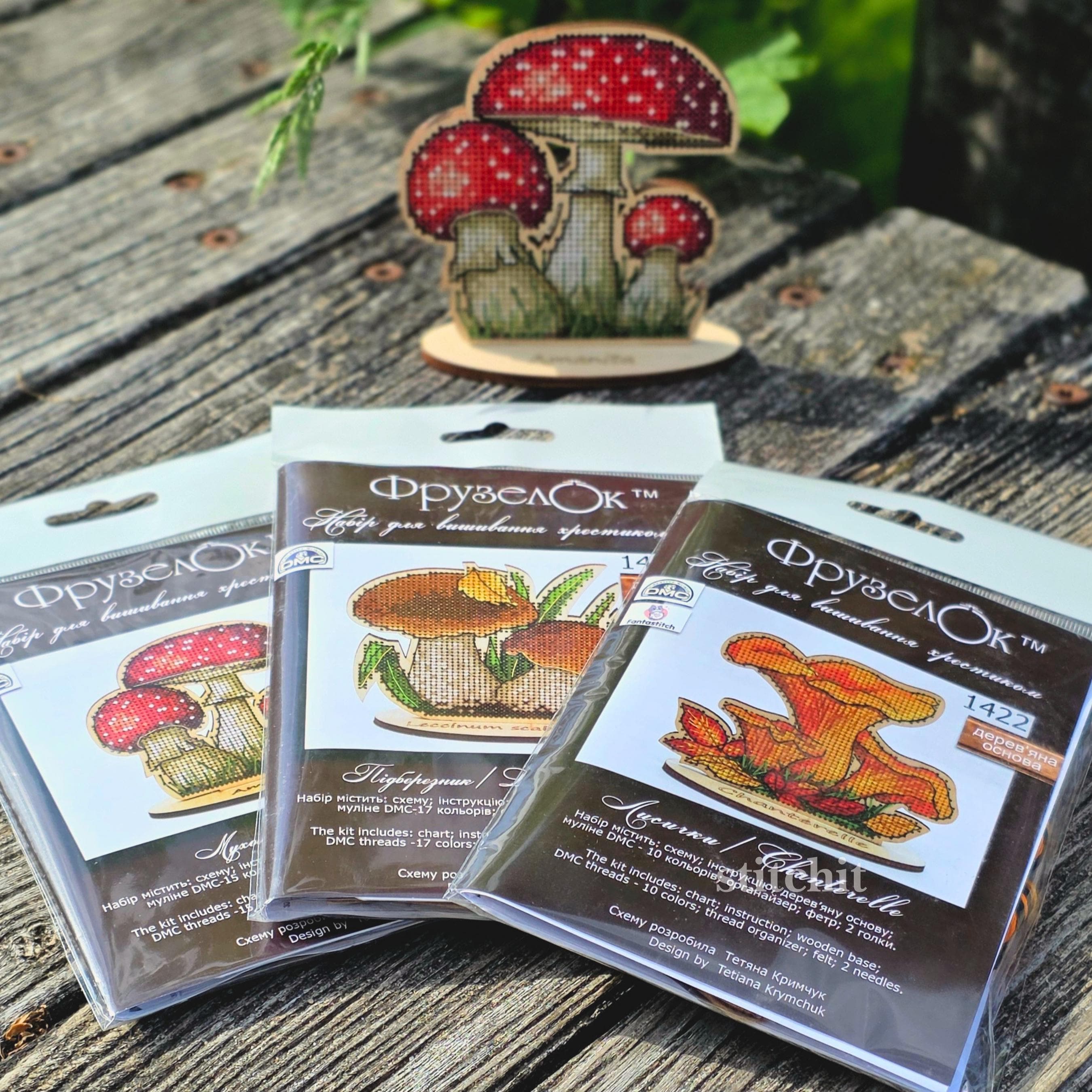 Chanterelle Cross Stitch Kit on Wood — three packaged mushroom kits beside a finished stitched mushroom on a wooden stand in an outdoor setting with greenery.