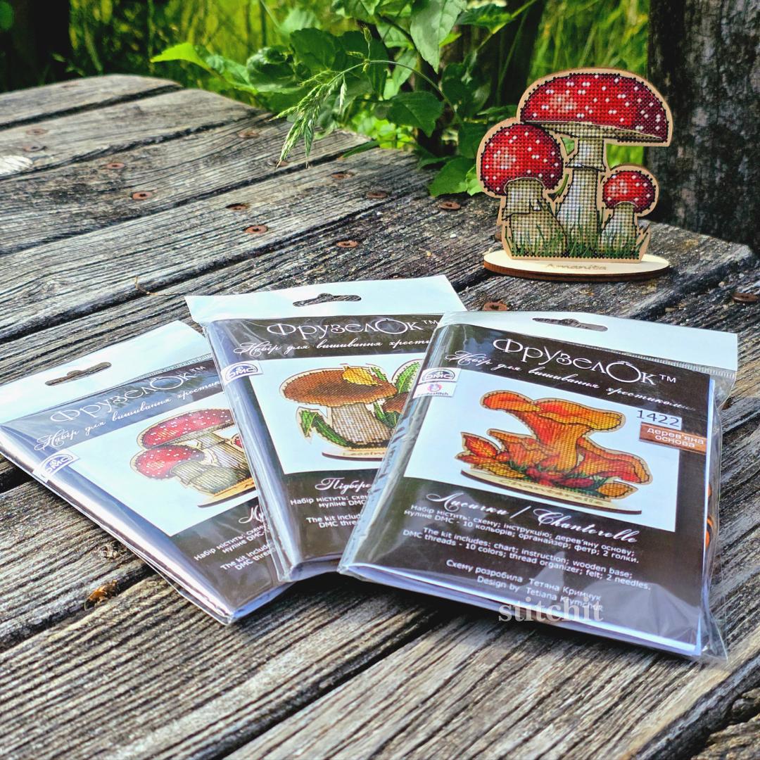 Mushroom Cross Stitch Kit on Wood — three packaged Amanita kits beside a finished stitched fly agaric on a wooden stand in an outdoor forest setting.