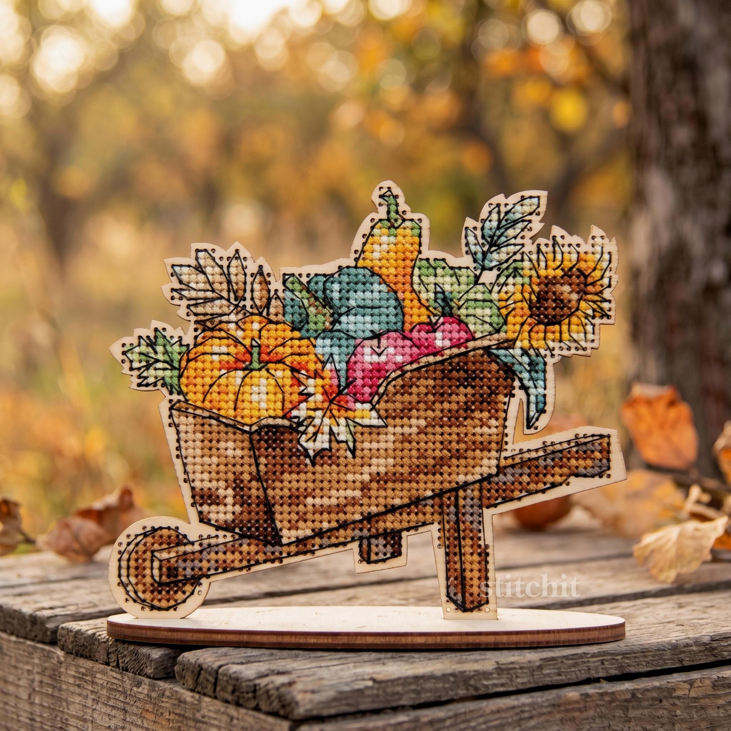 Harvest Cross Stitch Kit on Wood displayed in a wheelbarrow filled with vibrant autumn fruits and sunflowers.