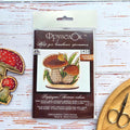 Forest Mushroom Cross Stitch Kit on Wood — Birch Bolete kit packaging beside two finished stitched mushrooms and scissors on a white wooden surface.Forest Mushroom Cross Stitch Kit on Wood — Birch Bolete kit packaging beside two finished stitched mushrooms and scissors on a white wooden surface